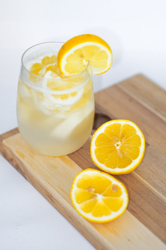 Icy cold glass of lemonade, adorned with sliced lemon wheels sitting on a wooden cutting board. Additional lemon slices are decoratively arranged.